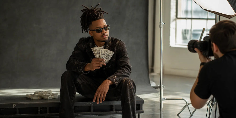 Music artist poses with prop money while a photographer shoots in a professional daylight studio.